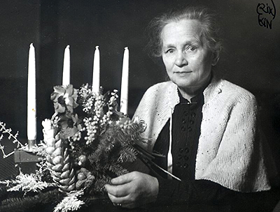 Foto av Agda Östlund som sitter vid ett bord där det står en adventsljusstake och något blomsterarrangemang. Agda har en ljus kofta på sig och ser in i kameran.