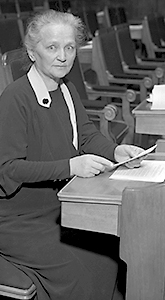 Foto av Agda Östlund sittande vid sin bänk i riksdagen. Hon håller i några papper och ser rakt in i kameran.