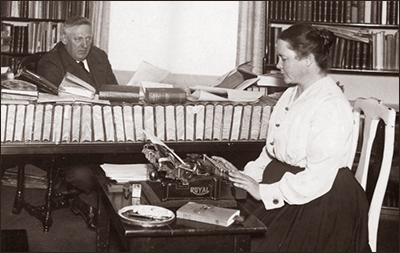 Foto av Maria Qvist som sitter och skriver på en skrivmaskin, bakom henne sitter Fabian Månsson med massor av böcker utslängda över bordet och en hel rad med böcker närmast Maria. I balgrunden syns bokhyllor