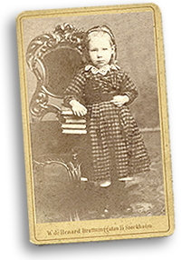 Maria Sandel på ateljébild som barn, iklädd rutig klänning med spetskrage och diadem i håret står hon lutad mot en stol med böcker på.