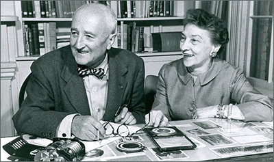 Foto av Elizebeth och William på äldre dar, de sitter vid ett bord där det ligger fullt med material, och i bakgrunden syns en bokhylla. Bägge ler stort och ser på något till vänster ur bild.