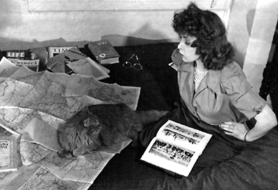 Foto av Peri-Purr och June Tarpé Mills som sitter på en säng eller soffa. Hon har en bok i knät och flera böcker samt ett par glasögon ligger i bakgrunden. Katten ligger på en stor utvecklad karta.