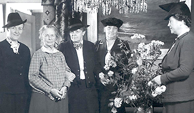 Foto av fem kvinnor. Alla utom Karolina Widerström är klädda i mörja dräkter och hatt. En stor blombukett står i förgunden. Nederdelen av en kristallkrona syns i toppen av bilden.