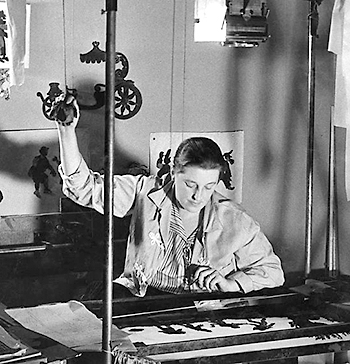 Svartvitt foto av Lotte Reiniger som sitter vid sitt kamerabord med siluetter. Hon har ena handen lyft och håller i ett snöte som verkar tillhöra maskinen/bordet. På väggen bakom henne syns fler siluetter.