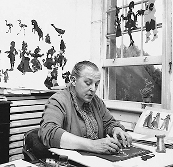 Foto av Lotte Reiniger sittande och tecknande vid ett bord. På väggen bakom henne och i fönstret händer färdigbyggda siluetter av människor och djur.