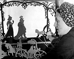 Svartvitt foto av Lotte Reiniger i profil intyill ett antal siluetter av vagnar med hästar med plymer och ovanför dem en prinsessa, en kung och en katt, omgivna av blad som slingrar sig längs med pelare.