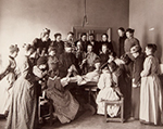 En mängd kvinnor i fotlånga klänningar är samlade runt ett bord där en manlig lärare håller på att dissekera ett lik. Bredvid läraren står ett skelett.