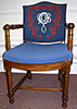 Foto av en gammaldags enkel stol med armstöd. Stolsistsen och ryggen är i blått tyg, och på stolsryggen är  institutets symbol broderad, den specialtillverkade broschen, här  omgiven av en slinga röda blad.