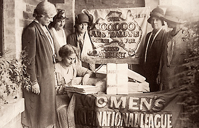 Foto av fem kvinnor stående runt en sittande kvinna som skriver på en tjock bunt papper. Längst fram en halv banderoll, det går att se texten "Women's International Leagur"