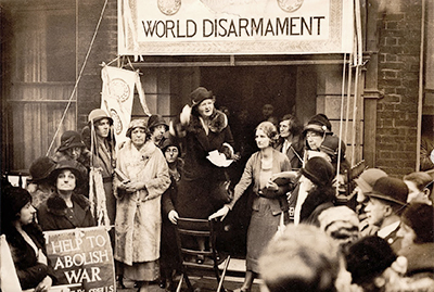 Foto av Margaret Bondfield som håller engagerat appelltal och gestikulerar med armarna, runt om henne står mestadels kvinnor och en del män