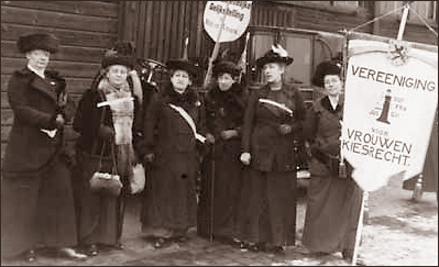 Foto av sex kvinnor utomhus. Det är nog kallt, för de har kappor och hattar på sig. En håller i ett standar med texten  Vereeniging voor Vrouwenkiesrecht