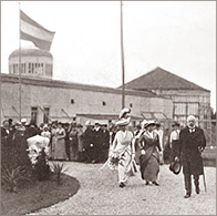 Foto av två kvinnor i hattar och långa kjolar som kommer gående på en grusplan med massor av folk stående bakom sig. Snett framför dem går en man med käpp och hatten i handen