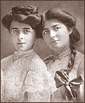 Foto av två unga tjejer, den ena med uppsatt hår, den andra mned en lång fläta med en rosett på. Båda ser in i kameran, den med uppsatt hår har en spetsklänning