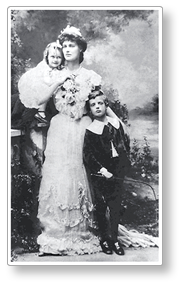 foto av Constance Makievics som står i en stilig vit klänning med dottern Maeve på ena armen och styvsonen Stanislas lutande mot henne på andra sidan