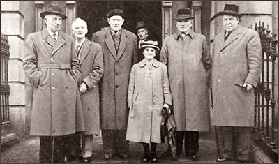 Foto av kortväxt Rosie Hackett i mitten, omgiven av tre män till vänster och två till höger. Alla männen är betydligt större än hon. De står framför en port på gatan med ytterkläder (rockar) på sig