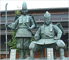 Foto av staty föreställande en stående Tomoe Gozen bredvid en sittande Yoshinaka. Hon håller sin naginata i ena handen.