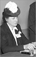 Foto i halvfigur av sittande kvinna iförd en hatt med plym och en ordförandeklubba och diverse papper framför sig
