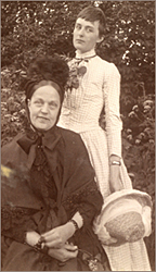 Foto av en sittande äldre kvinna i mörk klädsel och en ung kvinna i ljus klänning och hållande i en hatt, stående bakom den äldre.