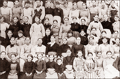 Foto av massor av kvinnor och flickor som sitter  i fyra rader bakom varandra iklädda tidstypiska långa kjolar och blusar