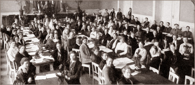 Foto av massor av kvinnor som sitter i en stor sal vid långa bord. Alla har papper framför sig på borden