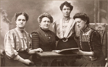 Foto av fyra kvinnor i kläder typiska för sekelskiftet runt 1900. Tre sitter vid ett bord, en står. En kvinna tittar i en bok, de andra ser in i kameran