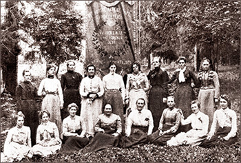 Foto av en grupp kvinnor i en skog, några sittande, andra stående. Bakom dem står ett stort standar, dubbelt så högt som någon kvinna.
