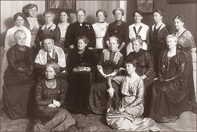 Foto av tio stående damer, sex sittande på stolar, och två sittande på golvet framflr de andra. De har typiska seklskifteskläder 1800/1900 på sig och är tydligt finare damer