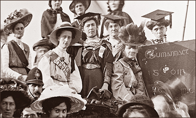Foto av ett antal kvinnor som står olika högt som på en kulle, men endast ansikten och halva kroppar syns. Till höger ett plakat med text på iriska. Till vänster syns ett huvud av en polis i kask mitt i.