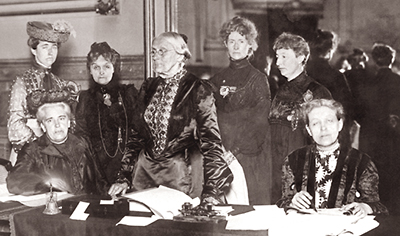 Foto av sju kvinnor i en sal vid ett bord, två sitter och fem står. I bakgrunden skymtar fler kvinnor.