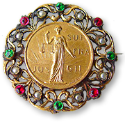 IWSA:s brosch med en inre ring av guld, där det står en kvinna med vågskål ioch texten JUS SUF FRA GII och runt den ett filigranarbete i guld med stenar i vitt, rött och grönt