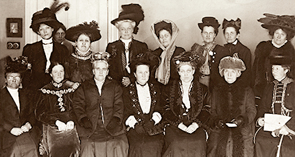 Foto av sju sittande kvinnor och åtta kvinnor som står bakom dem. De med högst hattar står bak.