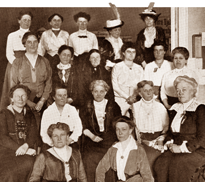 Foto av 18 kvinnor som sitter och står på fyra led. Två av dem som står längst bak har hattar med plymer på.