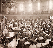 Foto av en jättesal med ljusiga lampor och proppat med folk som sitter och lyssnar. Till vänster står flera kvinnor på någon form av podium.