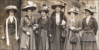 Sex kvinnor står i rad på gatan, vända mot kameran. Alla har dåtidens stora hattar och långa klänningar, men bilden är klippt runt knähöjd.