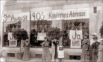 Foto av ett antal kvinnor på gatan utanför skyltfönster med norska flaggor och ordet "Ja" på affischer. Ovanför fönstren står: 13:de August 1905, Kvindernes Adresse
