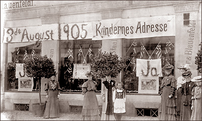 Foto av ett antal kvinnor på gatan utanför skyltfönster med norska flaggor och ordet "Ja" på affischer. Ovanför fönstren står: 13:de August 1905, Kvindernes Adresse