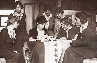 Foto av sju kvinnor som sitter eller står runt ett bord där en kvinna syr fast stjärnor på ett standar