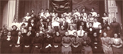 Foto av massor av kvinnor som sitter och står i rader inomhus. Längst bak står deras fana. Till höger syns en skylt med texten:  Läskedrycker och kaffe serveras, samt en hand som pekar åt höger