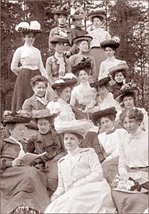 Foto av många kvinnor varav de flesta har stora hattar där de sitter eller står utomhus med skog i bakgrunden