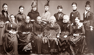 Foto av sju stående och sex sittande kvinnor i typiska 1800-talsklänningar. Flera har hatt. Alla ser in i kameran