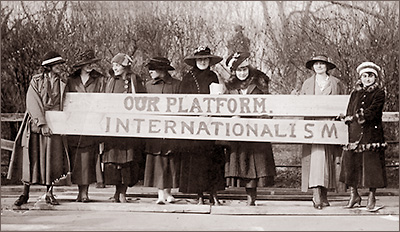 Foto av åtta kvinnor som står utomhus iförda hattar och kappor och håller upp vad som ser ut som två brädor med texten "Our platform" på den ena och "Internationalism" på den andra