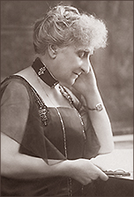 Halvfigurfoto av Ghénia Avril de Sainte-Croix sittande i profil med näsan åt höger. Hon ser ner i ejn bok hon har i handen på ett bord