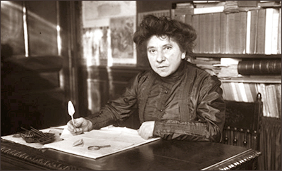 Foto av Hubertine Auclert sittande vid ett skrivbord med en penna i handen. Hon ser in i kameran