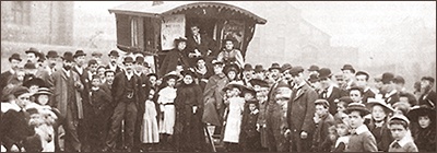 Foto av en vagn som det sitter två kvinnor och en man vid. Runt dem är en stor folksamling av både barn och vuxna.