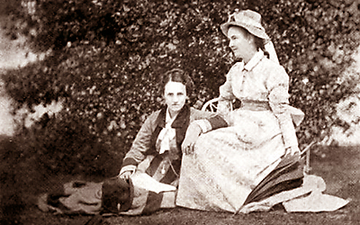 Foto av Rhoda och Agnes i deras trägård, Agnes - i vitt - sitter på en stol och Rhoda sitter på gräset nedanför henne och lutar ena armen i Agnes knä. Agnes har ett paraply i handen och en hatt som matchar klänningen. Rhoda ser rakt in i kameran