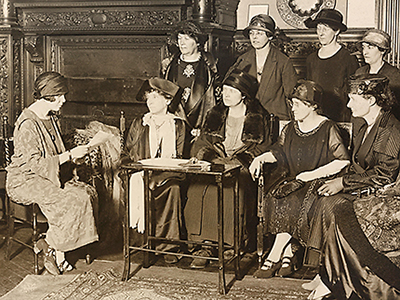 Foto av en kvinna till vänster och en grupp av 8 kvinnor till höger. Kvinnan till vänster läser upp något från ett papper hon håller i.