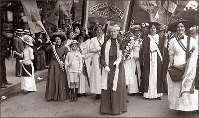 Foto av demonstrationståg där Charlotte Despard går längst fram. En fana med texten "Women's Freedom League" finns bakom henne. Till vänster säljer en kvinna en tidning till någon. Flera kvinnor bär flaggor.