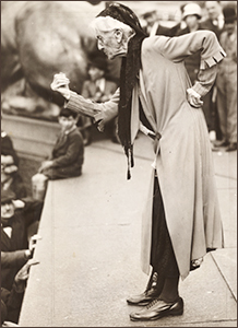 Foto av Charlotte Despard i helfigur. Hon står på en upphöjd del av Trafalger Square och talar till folk nedanför. Hon har knutit högerhanden och står böjd mot folket