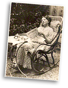Foto av Christabel sittande i en gungstol ute i en trädgård. Hon har en tidning i knät, men håller upp den så att titeln syns: Suffragette