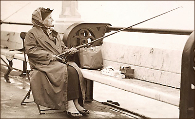 Foto av Christina Broome sittande på en hopfällbar stol framför en bänk. Hennes metspö vilar mot staketet, det ser ut att vara på en bro eller liknande. Hon har kappa och hatt och en sjal knuten över hatten runt hakan